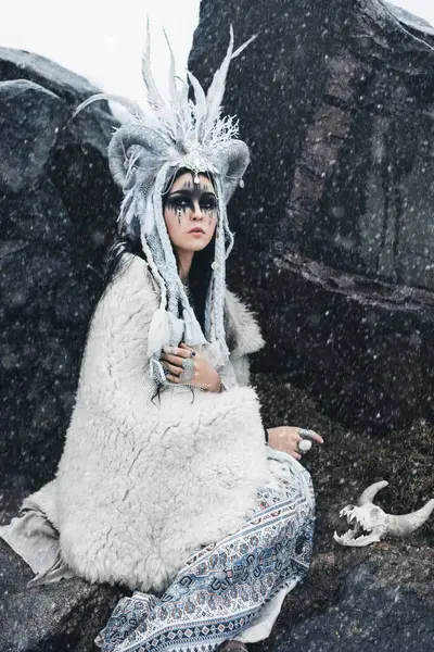 黒い髪とファンタジーメイクの美しい女性 ブレードと王冠で 角と羽 民族的なブーフスタイルの白い服で 秋の雪の中でリードに座って毛皮の帽子で — ストック写真