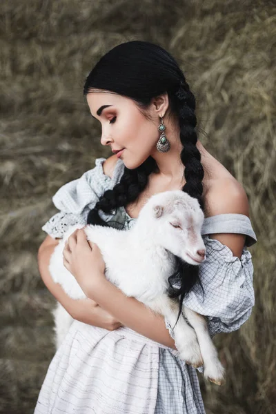 身穿白色民族服装的年轻貌美的黑发女人站在草垛里 怀里抱着一只山羊 奥托贝里节 — 图库照片