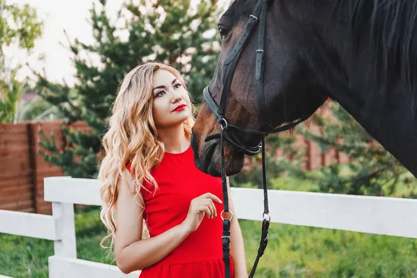 spring summer outdoor portrait of a beautiful blonde woman in a red long dress and with flowers in her hair standing in forest by the lake with a horse, hairstyle, makeup