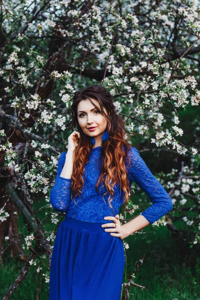 春天的户外肖像一个美丽的性感的黑发女人 卷曲的头发 蓝色的衣服和蓝色的耳环 苹果花 花园与树 — 图库照片