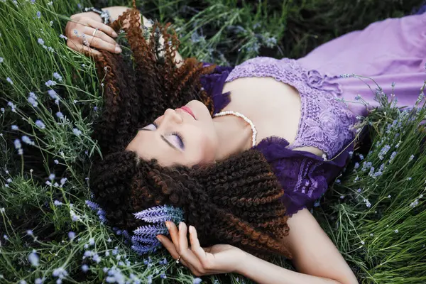 迷人的黑发女人 卷曲的头发 紫色的妆容 紫色的民族服装 头戴薰衣草花冠 躺在薰衣草花丛中的草地上 — 图库照片