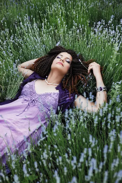 迷人的黑发女人 卷曲的头发 紫色的妆容 紫色的民族服装 头戴薰衣草花冠 躺在薰衣草花丛中的草地上 — 图库照片