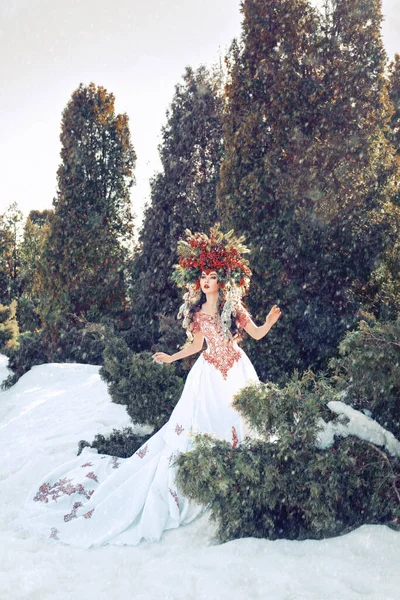 Fantasy winter queen standing in falling snow in the forest, a beautiful woman in the crown of red berries and branches