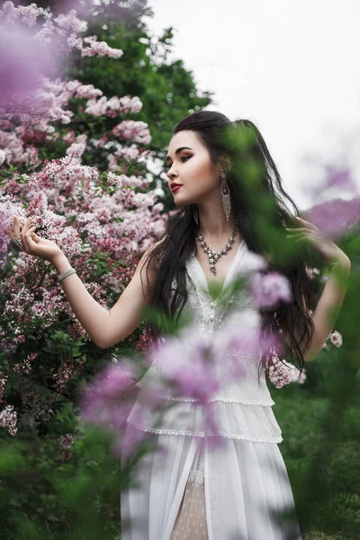 春天的户外画像 一个身穿白衣的漂亮的黑发女人站在紫丁香花丛中 — 图库照片