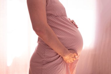 Beyaz hamile kadın pencerenin yanında karnına dokunuyor..