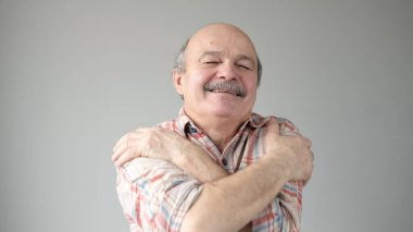 Kendine güvenen, gülümseyen, kendini kucaklayan yaşlı bir adam gri duvar arkasında tek başına. Olumlu insani duygular, yüz ifadesi. Kendini sev kavramı