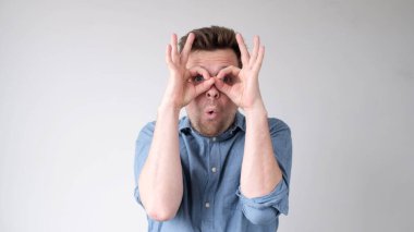 Genç bir Avrupalı adam sanki uzaktan bakıyormuş gibi parmaklarını dürbün gibi gözlerine tutar. Stüdyo çekimi