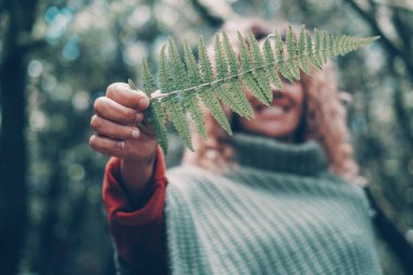 Büyük yeşil yaprak odaklı ön plan. Arka plandaki kadın gülümse ve büyük bir yaprak göster. Doğa ve çevre insanları aşk ve yaşam tarzı kavramını benimsiyor. Ormanların tahrip edilmesini durdurun. Açık hava parkında eğlence.