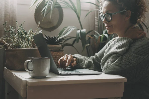 眼鏡をかけた幸せと穏やかな女性は自宅のコンピュータ上でウェブサーフィン 緑のムードカラー 背景に植物 屋内レジャーでラップトップで働く大人の女性 オンラインアクティビティ — ストック写真