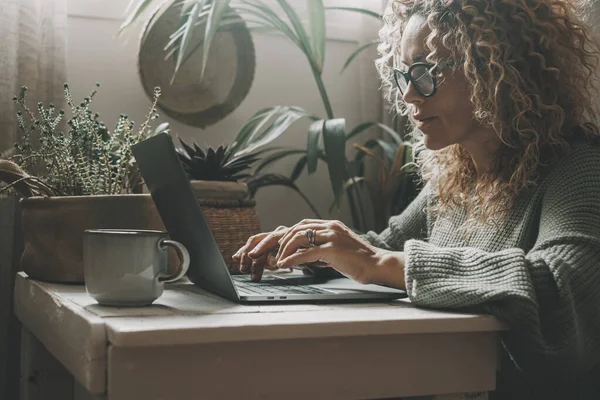 緑のムードカラーの背景と周囲のノートパソコンを使って家にいる女性です 背景に家の屋内庭園 魅力的な女性の人々は屋内レジャーライフスタイルで仕事やサーフィンのためのコンピュータを使用しています — ストック写真