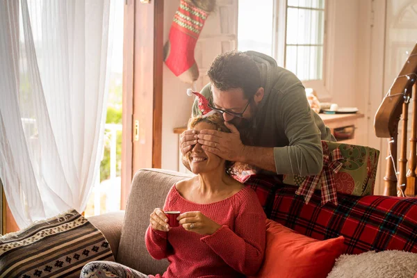 Happy couple enjoy Christmas holiday celebration at home with happiness. Man closing eyes to his woman to give her a xmas present gift. Surprise from behind concept. Traditional gift sharing leisure