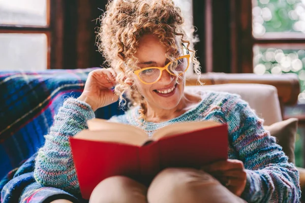 woman enjoying relax and indoor leisure activity at home reading a book with red cover. Education concept lifestyle. One happy female people read novel sitting at home. Smiling lady enjoying