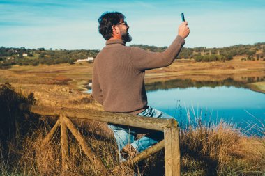  Erkek turist, göl manzaralı cep telefonuyla manzara fotoğrafı çekiyor. Parkta boş zamanların tadını çıkaran ve cep telefonuyla internet bağlantısı kuran bir adam var.