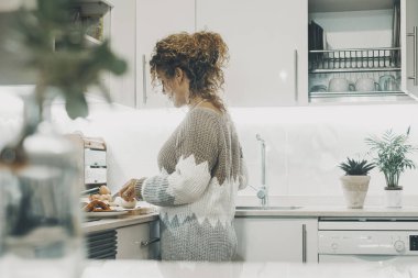 Beyaz, minimal, modern mutfakta yemek pişiren bir kadının yan görüntüsü. Bir kadın evde yemek pişiriyor. Zevk aktivitesi yalnız başına. Karım ya da bağımsız bir kadın içeride eğleniyor.