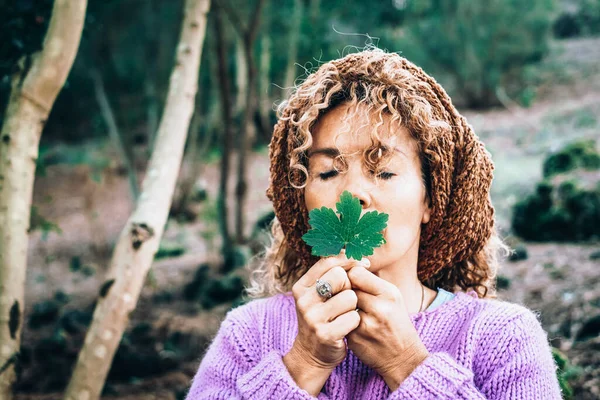 Nature and environment people concept lifestyle. One mature young woman holding a green leaf in front of her face and closing eyes to feel the nature outdoors. Alternative zen therapy for wellbeing