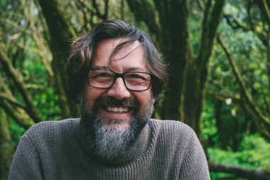 Arka planda yeşil orman ormanlarıyla gülümseyen ve kameraya bakan mutlu ve neşeli olgun bir adamın ön portresi. Parktaki insanlar doğanın tadını çıkarıyorlar. Çevre manzaralı bir yerde yetişkin bir erkek.