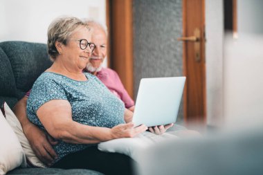 Senior couple at home having relax using laptop together. New modern lifestyle for mature retired people. Man and woman with computer and internet connection sitting on the sofa in indoor leisure