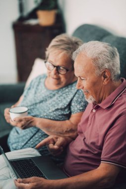 Old people using computer at home. Senior man and woman surfing the web on a wireless laptop connection. Modern retired lifestyle people enjoying notebook sitting and relaxing on the sofa together