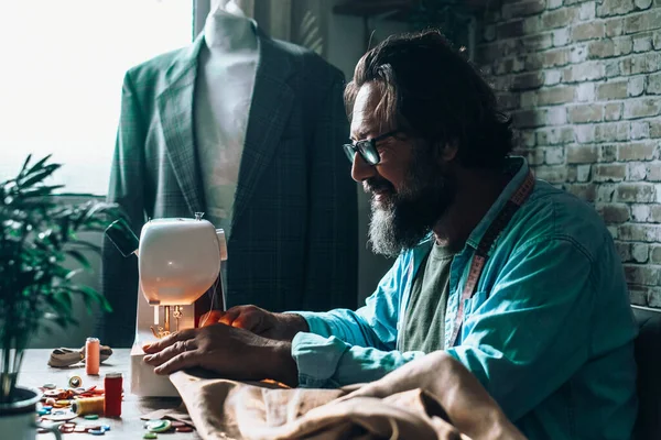 Tailor man working on sewing machine in workshop with dummy wearing suit in background. Mature happy man work indoor and smile. Concept of alternative job activity foe male people with beard
