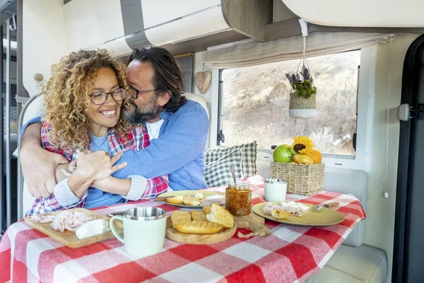 Excited and joyful couple in love inside camper van during lunchtime. Cheerful woman and man hugging and having fun laughing during adventure travel vacation on expedition vehicle transport. Love