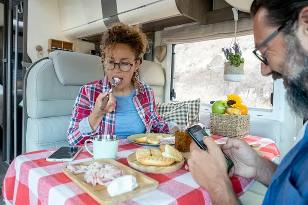 Man and woman eating lunch together using phone without talk to each other. Couple of modern traveler using connection roaming device mobile cell at the table. Camper van interior leisure activity people