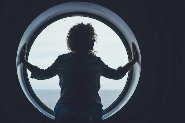 Portrait in dark shadow of a boat passenger sitting on a porthole window enjoying the trip. Travel people concept lifestyle. One female on cruise with seawater in background outside the window