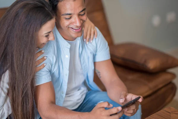 Young happy couple using mobile phone share social media news at home, smiling husband and wife millennial users customers talking going shopping online sit at table, mobile tech lifestyle concept