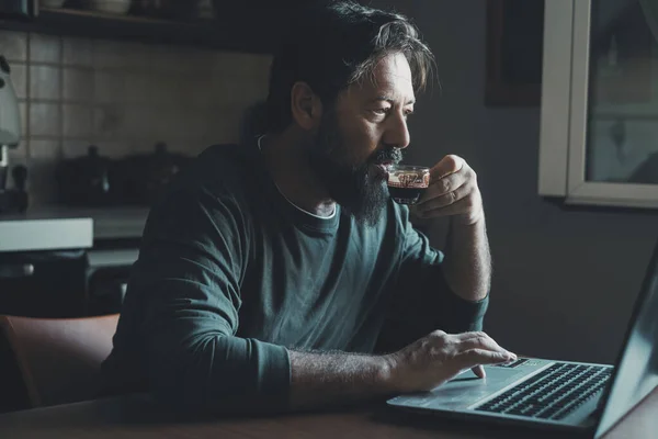 man alone at home using computer by night in the kitchen and drinking coffee to not fall asleep. Insomnia and healthy care. Male people overworking with laptop and online modern job. Stress Online