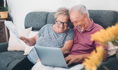 Aged couple at home paying bills online with laptop and laughing a lot of having fun together. Happiness and elderly lifestyle. Man and woman, old senior sitting on sofa with computer online connection