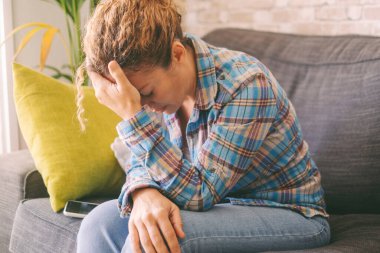  Exhausted and stressed woman at home sitting on the sofa touching her head, suffering. Sadness and depression, people disease. Loneliness. Female crying alone. Domestic problems couple concept