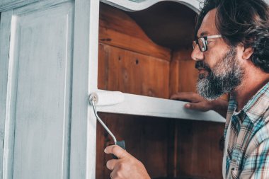 Close up of mature man painting and restoring an old antique wooden cabinet at home with roller and white paint. Recycling and upcycling, oldies. People working in leisure hobby activity alone