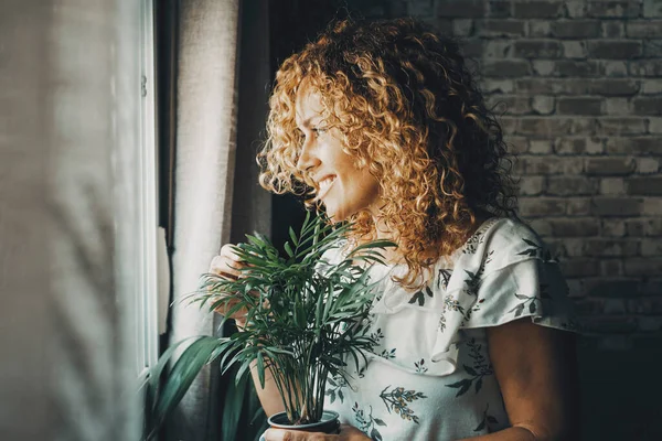 Happy woman at home looking outside the window and smiling inside dark light . Holding plant. Daydreaming concept lifestyle, female people. Good feeling young lady inside home leisure. Emotions