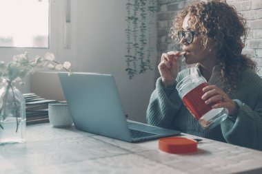  Evde dizüstü bilgisayar kullanan ve kutudan kurabiye yiyen bir kadın. Kötü beslenme yiyeceği kavramı kadın yaşam tarzını ve yerleşik iş aktivitelerini etkiler. Şirin bayan evde tek başına sörf yaparken eğleniyor.