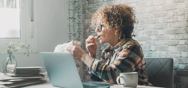 テーブルの女性は休憩中にチョコレートクッキーを食べてる ラップトップで働く女性や家庭のオフィスで食事をする女性 現代人のビジネスライフスタイルスマートジョブオンライン 起業家 — ストック写真