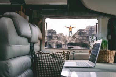 Dijital göçebe insan hayatı ve açık hava özgürlüğüyle alternatif ofis. Laptop karavanın içinde. Adam dışarıda bir kayanın üzerinde mutlu ve özgür duruyor. Uzaktan işçi dışarıda mutluluğun tadını çıkarıyor.