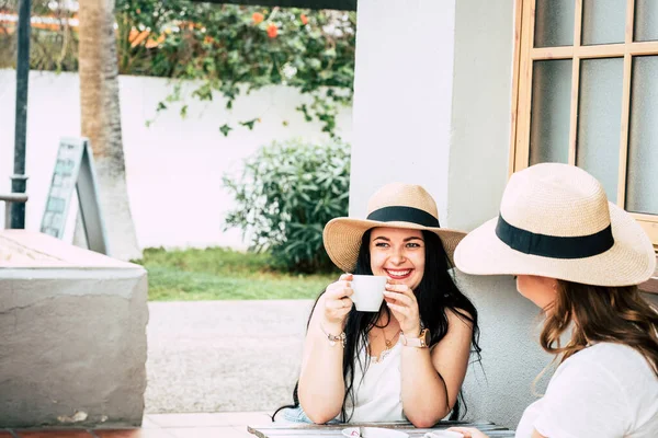 Young woman taking coffee with friend and smile. Happy tourist people in outdoor leisure activity drinking cappuccino in friendship. Girls together having fun wearing straw hats.
