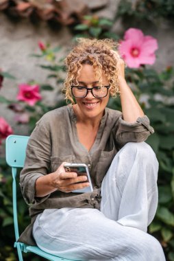  Kadın cep telefonu kullanıyor ve bahçede tek başına bir sandalyede oturup rahatlıyor. Cep telefonundaki kadın yazısının neşeli portresi. Görüntülü arama yapan bir bayan. Genç, olgun insanlar dinleniyor.