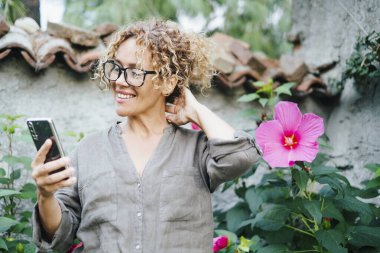 İnsanlar dışarıdaki teknoloji aktivitelerinde cep telefonu iletişimini kullanıyorlar. Arkadaşlarıyla görüntülü konuşmada gülümseyen kadın. Bahçede bitkiler ve çiçekler var. Neşeli bayan cep telefonundan arıyor.