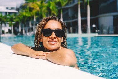 Mutlu kadın otel ve tatil yerlerinde güneşlenmeyi ve havuzu seviyor. Yaz tatilindeki insanlar. Seyahat ve lüks yaşam tarzı. Havuz kenarında gülümseyen ve mavi suda tek başına güneş gözlüğü takan genç bir bayan.
