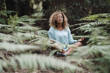 Çevresi yeşil tropikal yapraklarla çevrili orman parkında meditasyon yapan bir kadın. Yoga pozisyonu almış sağlıklı kadınlar ormanda yerde oturuyorlar. İyi olmak.
