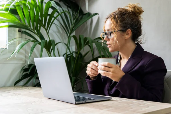 自宅のオフィスのデスクでノートパソコンの前で休憩コーヒーを飲みながらエレガントなスーツを着た女性マネージャー 現代の女性が働いていて 眼鏡をかけて画面を読んでいます フリーランス — ストック写真
