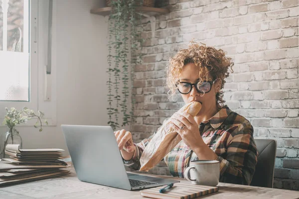 机の上のコンピューターで働いてる女が仕事を止めずに昼休みに大きなサンドイッチを食べてた ビジネスの人と食事の時間 フリーランスのスマートワークライフスタイル 現代の新しい仕事のライフスタイル — ストック写真