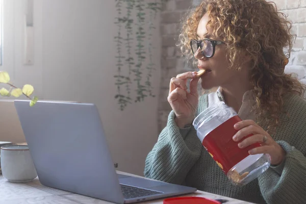 テーブルの上にノートパソコンを使い箱からクッキーを食べる女性です ゴミを食べながら仕事をする人たち 朝食や休憩中にコンピュータで働く忙しい人 現代の生活 — ストック写真