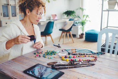 Boş zaman aktivitesi konsepti olan insanlar. Oturma odasındaki masada üzerinde tablet cihazıyla boncuk kolye yapan kadın. Modern alternatif yeni iş çalışmaları İnternet 'te satılıyor