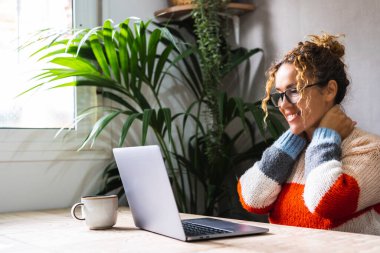 Genç girişimci dinlenmek için ara veriyor ve dizüstü bilgisayarın içeriğini izlerken gülümseyerek boynuna dokunuyor. Çevrimiçi modern iş. Evdeki bilgisayarları kullanan insanlar internette tek başlarına mutlu mesut dolaşıyorlar.