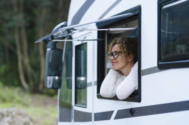 Minibüs turisti dışarıda keyfine bakıyor, arabanın camında dinleniyor. Karavan ve vanlife yaşam tarzıyla alternatif tatil kavramı. Kadınlar gülümser ve sakin bir ifade takınırlar.
