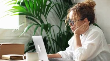 Modern kadın girişimci dizüstü bilgisayarda sıkıcı işler yapıyor. Çevrimiçi iş ve can sıkıntısı. İnsanlar kapalı ofis işyerinde çalışıyor. Mutlu değiller. Bitkili defter kullanan yorgun ve stresli bir bayan. 