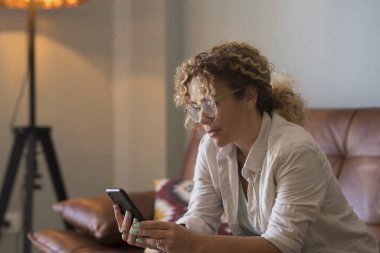 Evde tek başına oturan bir kadın oturma odasındaki koltukta oturan cep telefonu uygulamasını kullanmaktan sıkılmış. Kapalı alan teknolojisi iletişim eğlence aktivitesi. Yetişkin bir kadın cep telefonu içeriğine bakıyor.