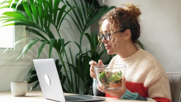 自然な健康的なライフスタイルの食べ物でランチをする準備ができてテーブルの上のサラダの緑のボウルでラップトップに取り組むかなり大人の女性 女性は忙しいコンピュータで働き メガネをかけています — ストック動画
