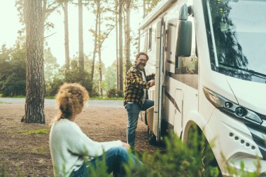 Yetişkin çiftler, modern bir karavan karavanı dışında birlikte boş vakit geçirmeyi severler. Seyahat eden insanlar tatil yaşam tarzı. Arka planda ağaçlar ve ormanlar olan doğanın ortasına park etmek. Özgürlük
